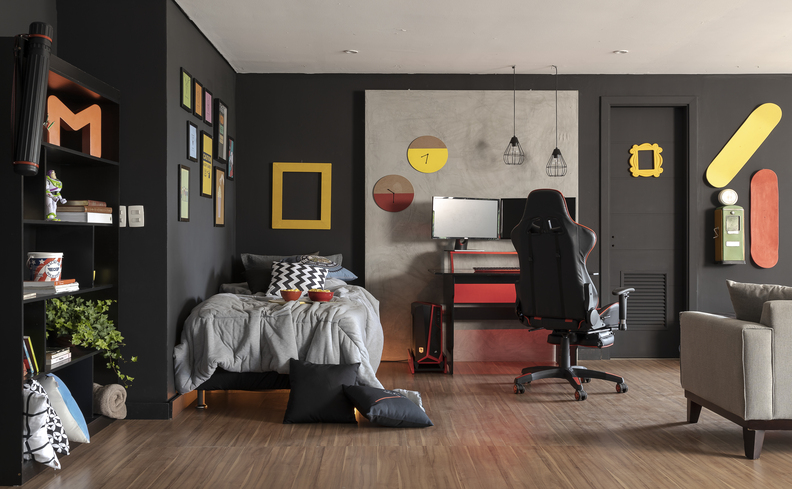 home office pequeno quarto com escrivaninha pequena, cadeira gamer, luminárias penduradas no teto, vários quadros decorativos e itens de escritório.