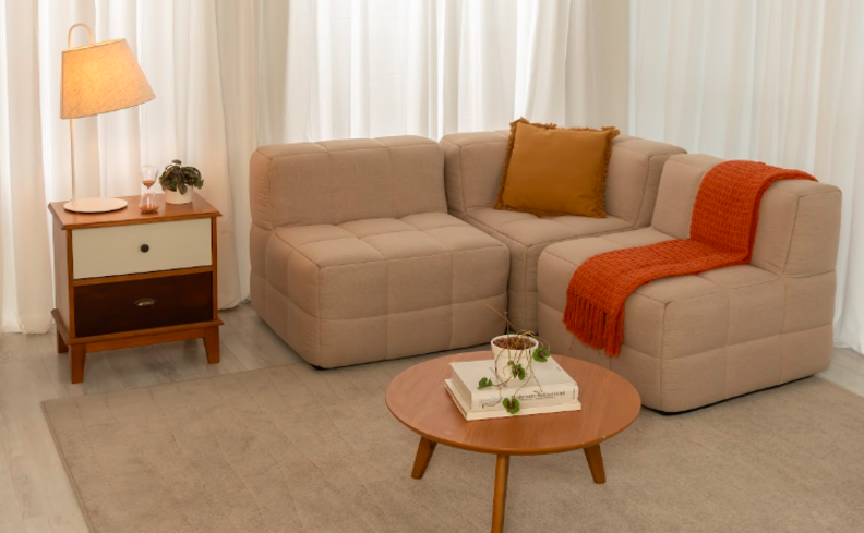 mantas de trico para sofá bege na sala de estar, com manta laranja, tapete bege, mesa lateral e mesa de centro, além de luminária de mesa e almofada.