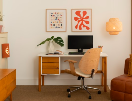 mesa para home office com escrivaninha de madeira com duas gavetas, quadros decorativos, luminária de teto, cadeira de escritório tudo em uma sala de estar.