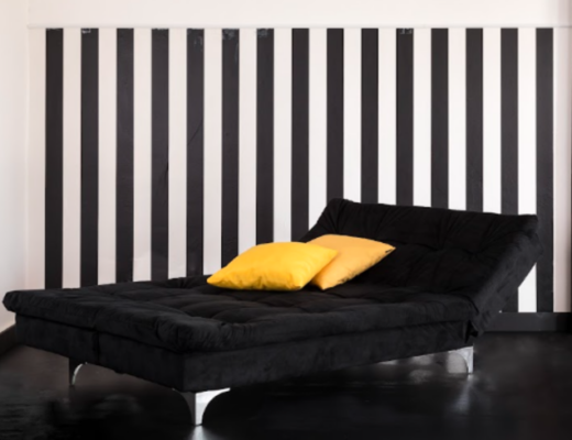 modelos de sofá-cama em uma sala de estar com um modelo na cor preta, duas almofadas amarelas e a parede com papel de parede listrado em preto e branco.