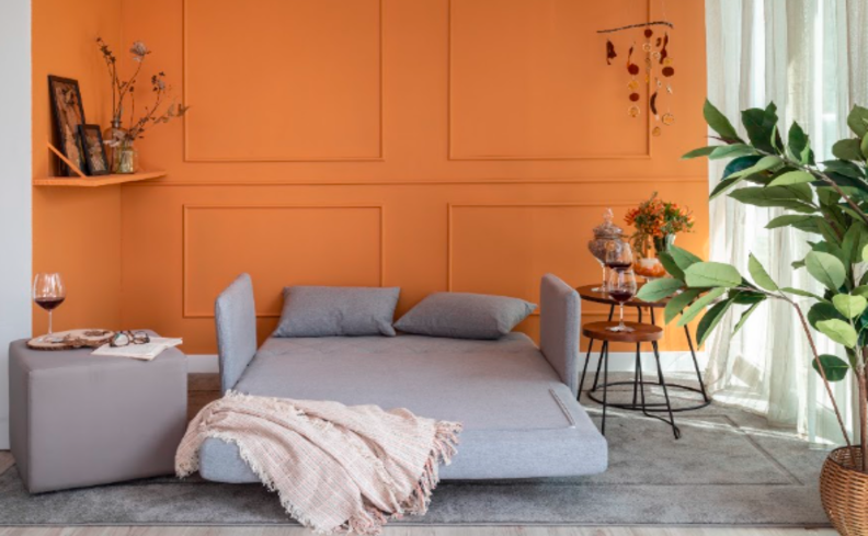 modelos de sofá-cama na cor cinza, totalmente aberto na sala de estar, com manta para sofá, vaso com planta, mesa de apoio e estante com diversos itens decorativos.