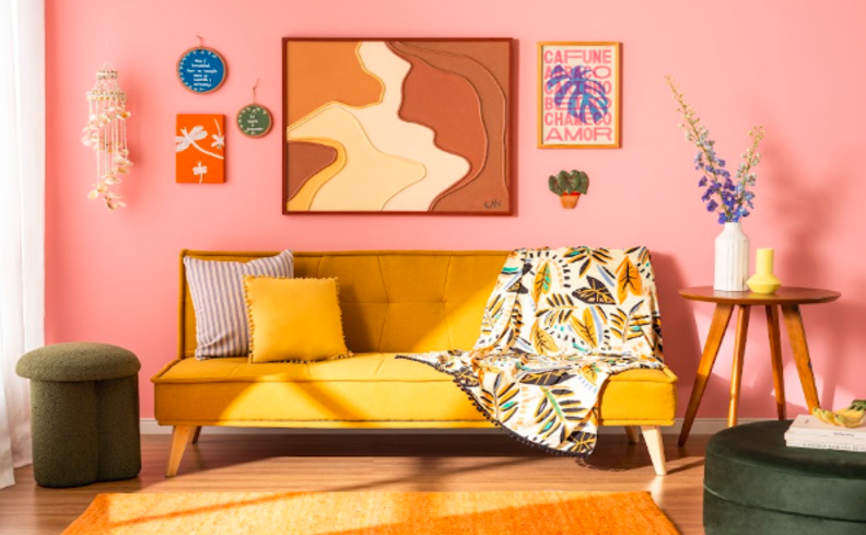 sofá-cama 2 lugares na cor amarela na sala de estar com parede rosa, quadros decorativos, pufe, mesa de apoio, manta para sofá e duas almofadas.