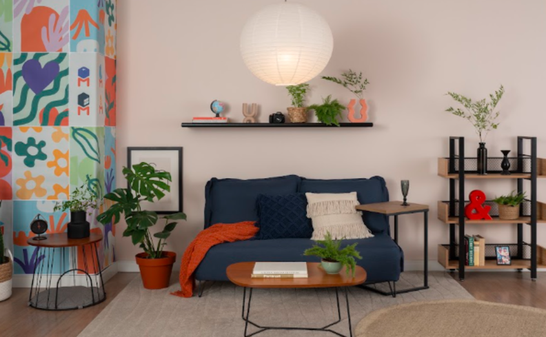 sofá-cama retrátil na cor azul, mesa de centro em madeira e aço, parede branca e metade colorida, estante de livro, vaso com planta, prateleira com diversos itens decorativos e almofadas.