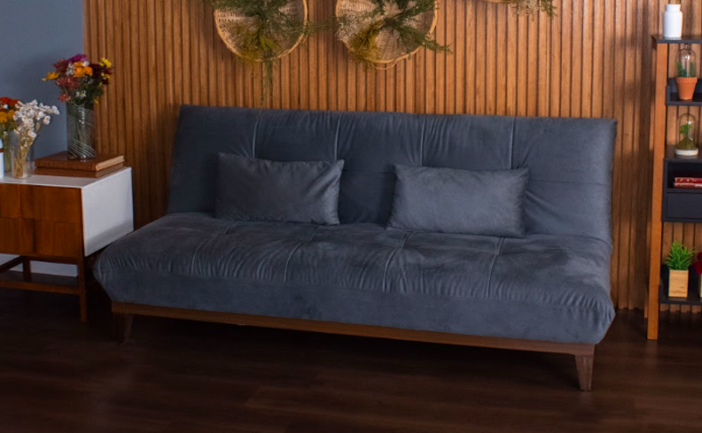 sofá-cama de 2 lugares na cor azul, com duas almofadas da mesma cor, parede em madeira ripada, estante de madeira, mesa lateral em tom de madeira e branco na sala de estar.